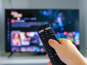 Ręka z pilotem z przyciskiem Netfilxa. W tle telewizor