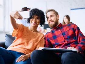 Uśmiechnięta para robi sobie selfie telefonem, w tle inni ludzie w biurze