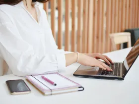 Kobieta w białej koszuli siedzi przy biurku i pisze na laptopie; obok telefon