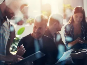 Grupa osób siedzi na spotkaniu, jeden mężczyzna uśmiecha się, trzymając wykres