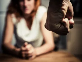 Na pierwszym planie ściśnięta w pięść dłoń, w tle widać przestraszoną kobietę
