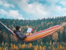 Mężczyzna leżący w hamaku czyta książkę, w tle znajdują się góry