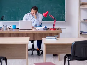Nauczyciel w klasie, ze zmęczenia masuje skronie, siedząc przy biurku z laptopem